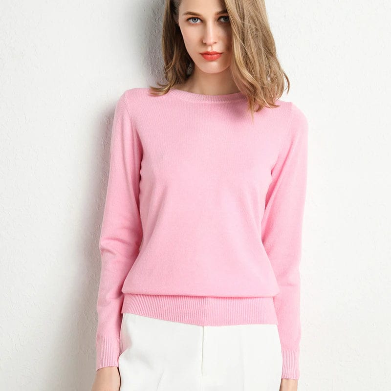Femme portant un pull rose en cachemire, manches longues, col rond, posant contre un mur blanc. Mode féminine élégante, tenue automne-hiver.