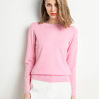 Femme portant un pull rose en cachemire, manches longues, col rond, posant contre un mur blanc. Mode féminine élégante, tenue automne-hiver.