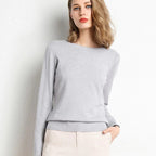 Jeune femme portant un pull gris élégant, manches longues, col rond, posant contre un mur blanc. Mode féminine, style décontracté chic.
