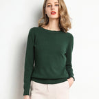 Jeune femme portant un pull vert élégant, manches longues, posant contre un mur blanc. Mode féminine, style automne-hiver, tenue décontractée.