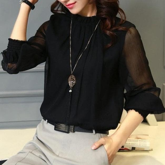 Femme portant un chemisier noir élégant avec manches transparentes, collier pendentif feuille, assise, style chic et moderne, mode féminine.