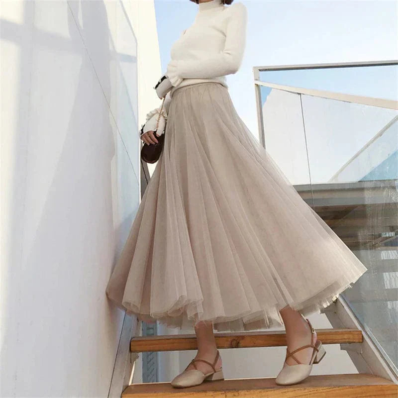 Femme en jupe longue beige fluide, haut blanc, escaliers en bois, style élégant, mode automne, chaussures à talons, lumière naturelle.