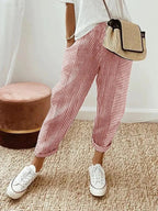 Femme portant un pantalon rayé rouge et blanc, t-shirt blanc, baskets blanches, sac en osier, debout sur un tapis beige. Mode décontractée chic.