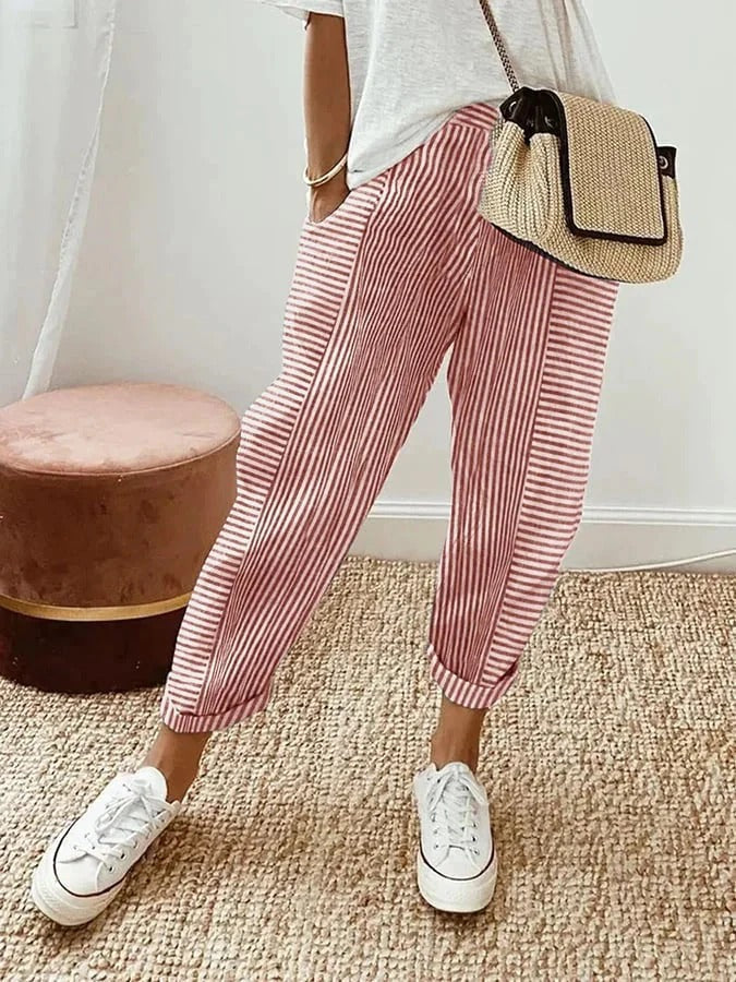Femme portant un pantalon rayé rouge et blanc, t-shirt blanc, baskets blanches, sac en osier, debout sur un tapis beige. Mode décontractée chic.