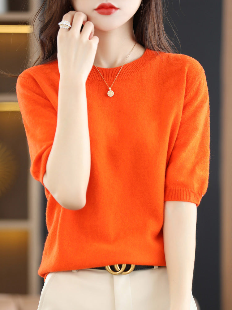 Femme portant un pull orange vif, manches courtes, avec un collier pendentif et une ceinture noire. Mode élégante, tendance automne-hiver.