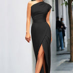 Robe noire élégante asymétrique avec fente, portée par une femme en extérieur urbain. Mode chic, tendance soirée, style moderne.