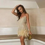 Jeune femme en robe à franges dorées et plumes, posant élégamment dans un intérieur moderne, tenant une pochette dorée. Mode soirée chic.