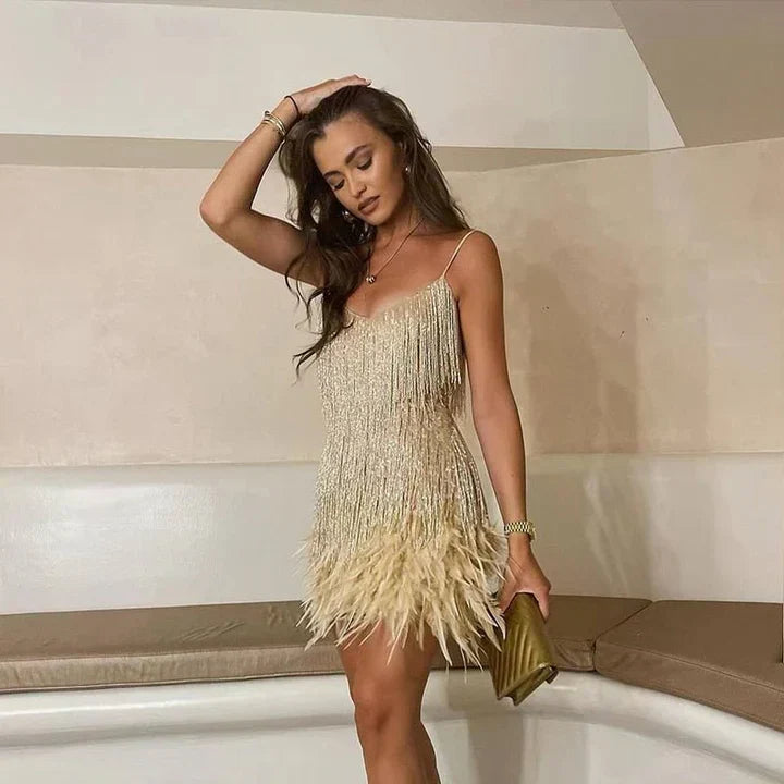 Jeune femme en robe à franges dorées et plumes, posant élégamment dans un intérieur moderne, tenant une pochette dorée. Mode soirée chic.
