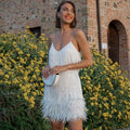Femme souriante en robe à franges blanches, tenant une pochette, devant un mur de briques et des fleurs jaunes. Mode élégante, soirée d'été.
