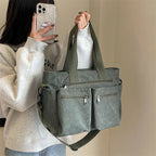 Sac à main en toile vert olive avec poches zippées, porté par une personne en sweat gris. Accessoire mode tendance, pratique et spacieux.