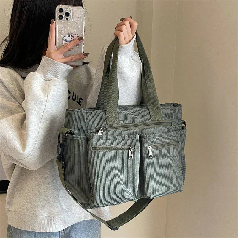 Sac à main en toile vert olive avec poches zippées, porté par une personne en sweat gris. Accessoire mode tendance, pratique et spacieux.