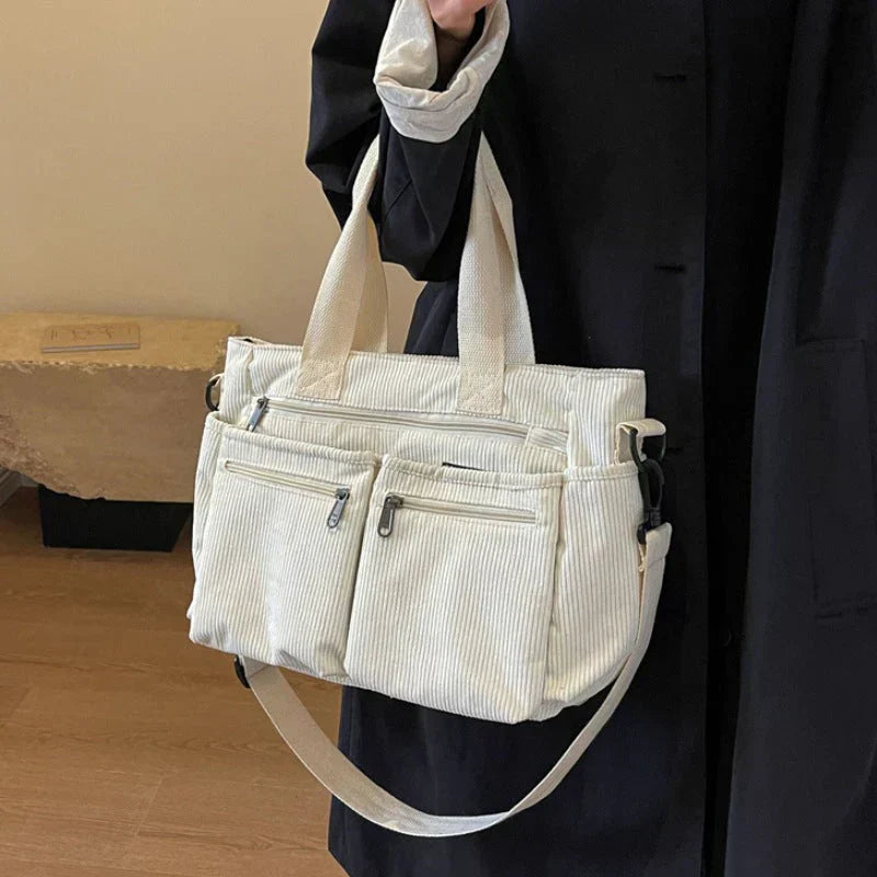 Sac à main en toile blanche avec plusieurs poches zippées, porté par une personne en manteau noir. Accessoire mode pratique et élégant.