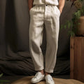 Pantalon en lin beige pour homme, style décontracté, avec ceinture, associé à des baskets blanches, ambiance naturelle avec plantes en arrière-plan.