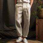 Pantalon en lin beige pour homme, style décontracté, avec ceinture, associé à des baskets blanches, ambiance naturelle avec plantes en arrière-plan.