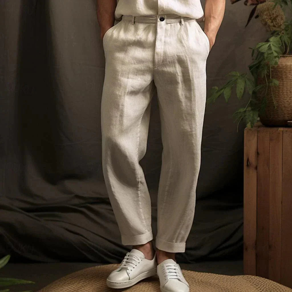 Pantalon en lin beige pour homme, style décontracté, avec ceinture, associé à des baskets blanches, ambiance naturelle avec plantes en arrière-plan.