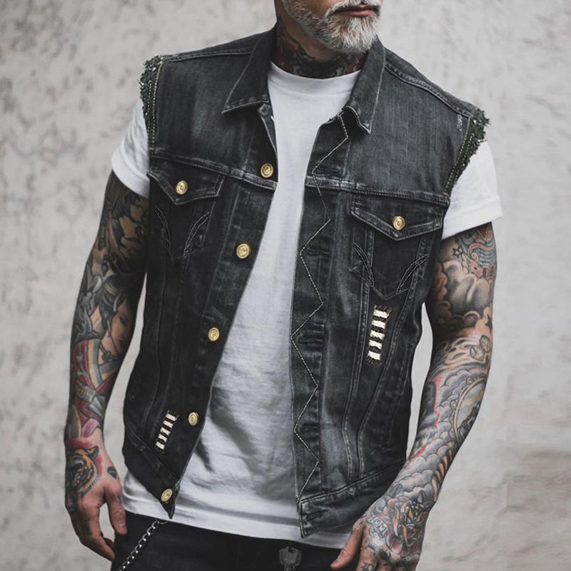 Homme tatoué portant un gilet en jean noir avec boutons dorés, sur un t-shirt blanc. Mode urbaine, style décontracté, tendance vestimentaire.