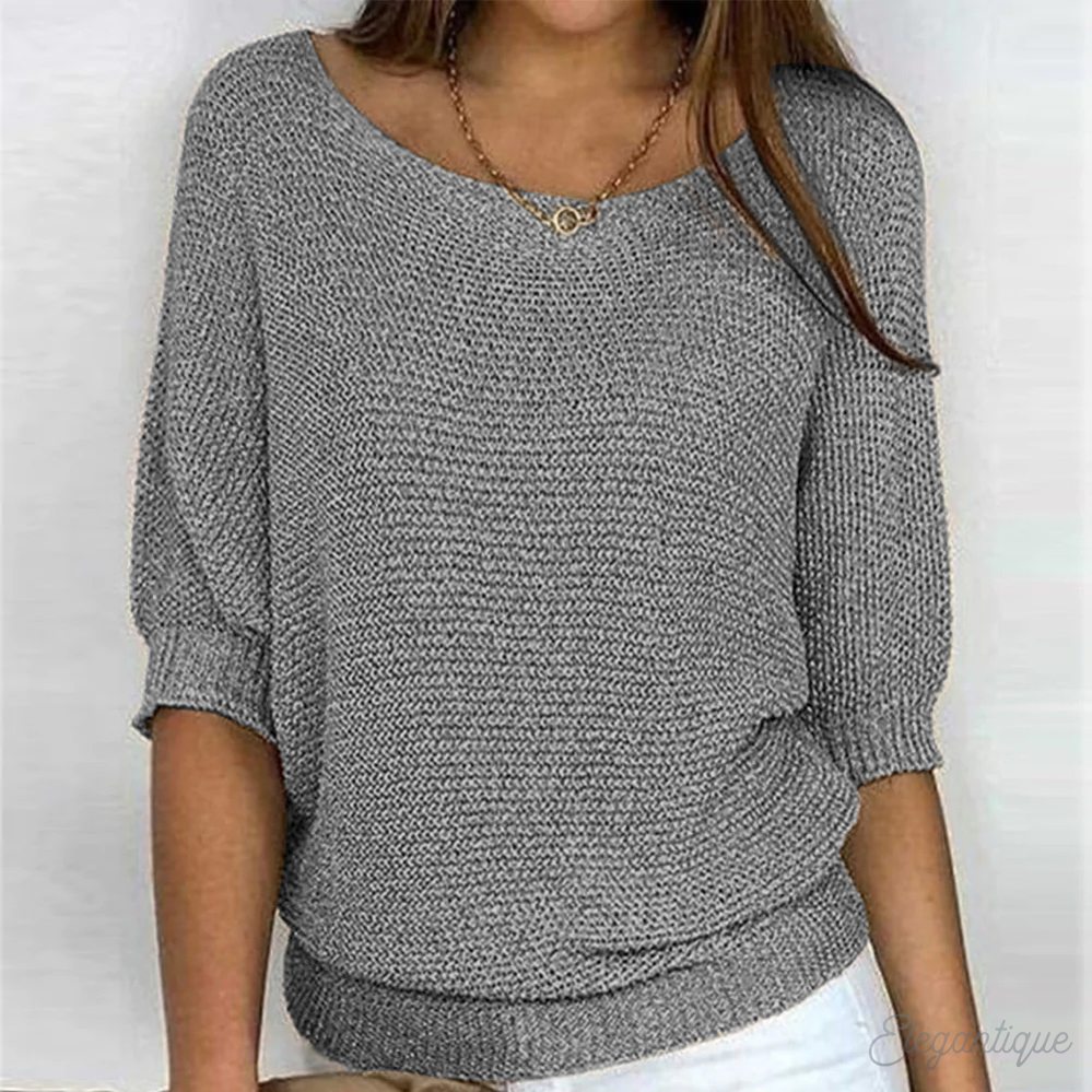 Femme portant un pull gris en tricot, manches trois-quarts, col bateau, avec un collier doré, sur fond blanc. Mode féminine élégante et décontractée.