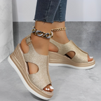 Sandales compensées dorées brillantes pour femme, avec semelle blanche épaisse, portées avec un jean ajusté et bracelet chaîne doré.