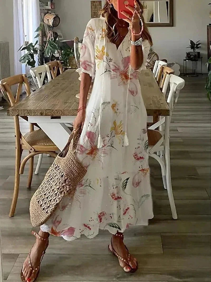 Robe longue blanche à fleurs portée par une femme, avec un sac en osier, sandales marron, dans une salle à manger moderne. Mode estivale élégante.