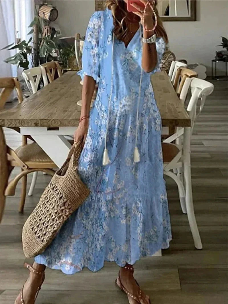 Robe longue bleue à fleurs portée par une femme, avec un sac en osier, dans une salle à manger moderne. Mode estivale, style bohème chic.
