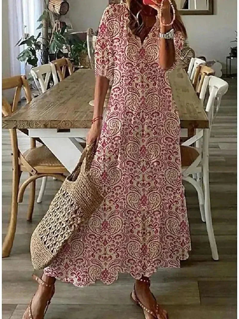 Robe longue bohème à motifs floraux rouges et blancs, femme tenant un sac en osier, sandales marron, intérieur avec table en bois et chaises.