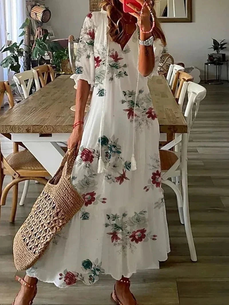 Robe longue bohème blanche à motifs floraux rouges et verts, femme tenant un sac en osier, intérieur moderne avec table en bois et chaises.