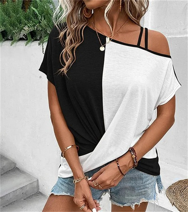 Femme portant un t-shirt bicolore noir et blanc, épaules dénudées, avec des bijoux dorés et un short en jean, posant devant un mur blanc.