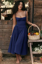 Jeune femme en robe bleue élégante, posant pieds nus près d'un panier de légumes colorés, devant une cabane en bois rustique.