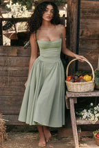 Jeune femme en robe verte élégante, posant avec un panier de légumes frais devant une cabane en bois rustique, ambiance champêtre et naturelle.