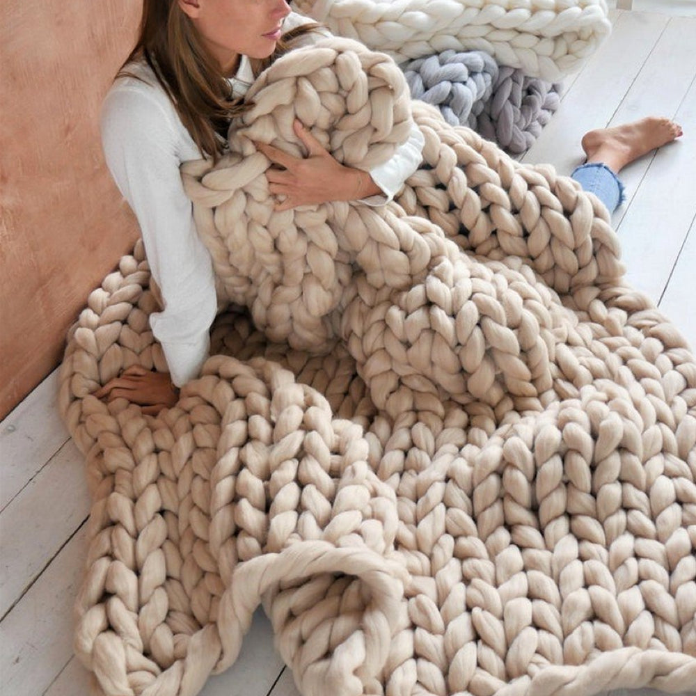 Femme assise avec un plaid en laine épaisse tricotée à la main, couleur beige, sur un sol en bois blanc. Couverture cosy, décoration intérieure moderne.