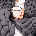 Femme tenant une tasse blanche émaillée, enveloppée dans une couverture en laine épaisse grise. Confort, chaleur, détente, hygge, cocooning.