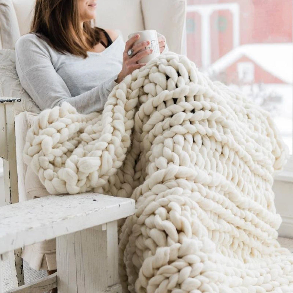 Femme relaxant sous une couverture en laine épaisse, tenant une tasse, assise près d'une fenêtre enneigée. Confort, hiver, détente à la maison.