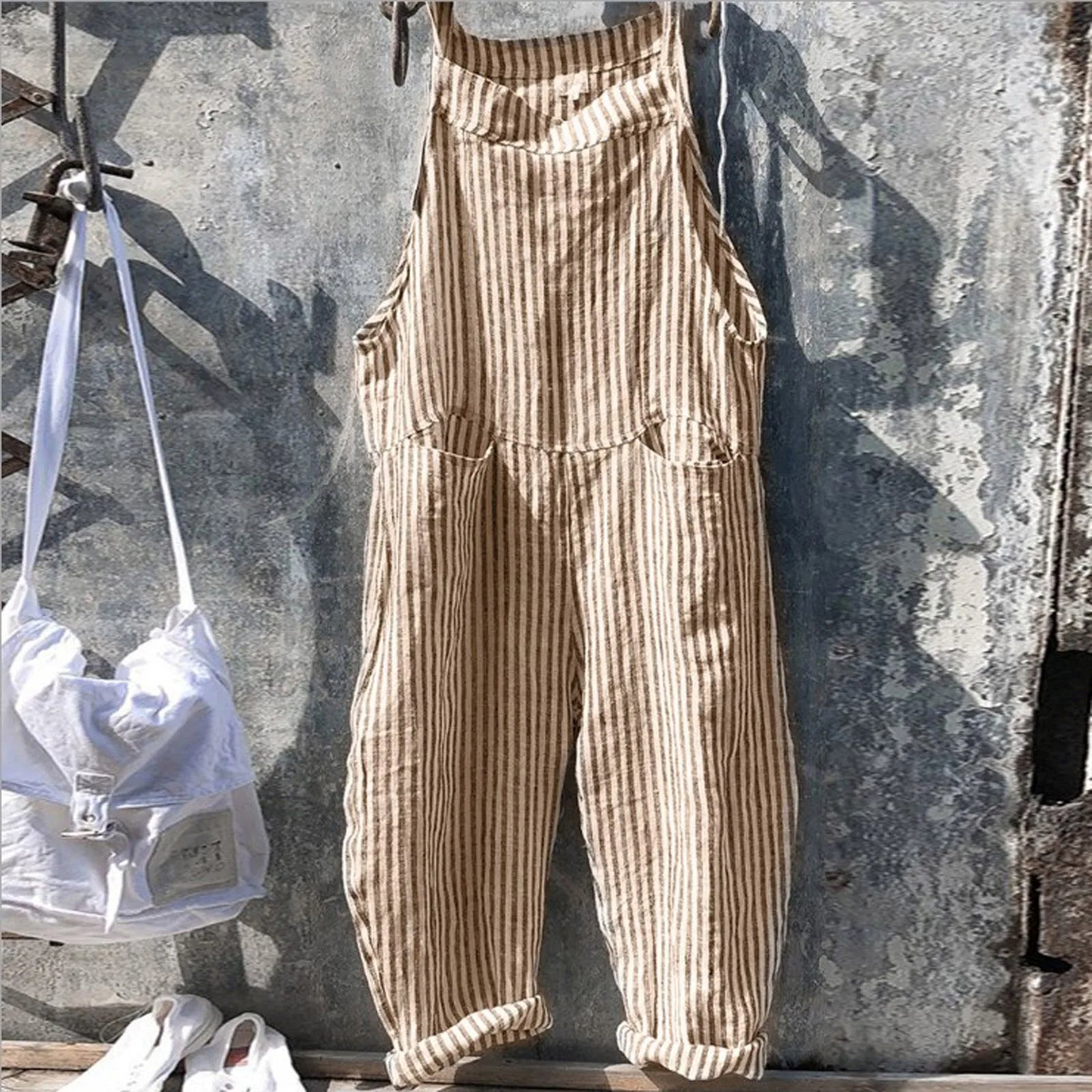Salopette rayée beige suspendue sur un mur texturé, accompagnée d'un sac blanc et de chaussures blanches. Mode décontractée, style vintage.