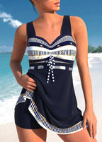 Femme en maillot de bain bleu marine à rayures blanches et dorées, sur une plage ensoleillée avec mer turquoise en arrière-plan. Mode plage élégante.